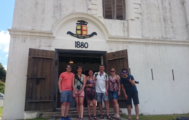 a group of tourists pose for a picture in front of a colonial style building on their Kuching day tour.
