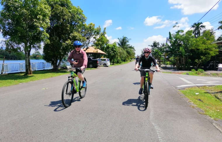 A couple cycle along a road on a sunny day on their Kuching day tour.