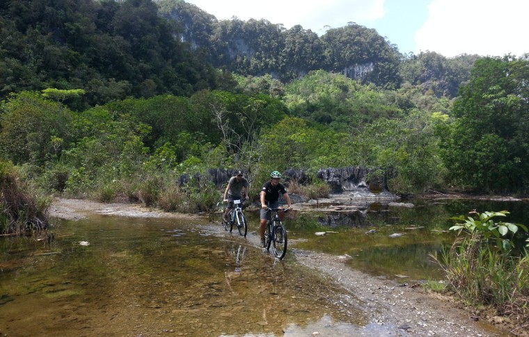 A couple cycle over a trail through a tranquil river on their Fairy Cave Kuching tour.