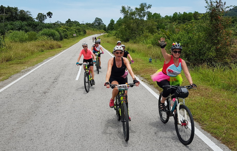 Guests smile at the camera as they cycle along a road surrounded by greenery on their Borneo cycling tour.