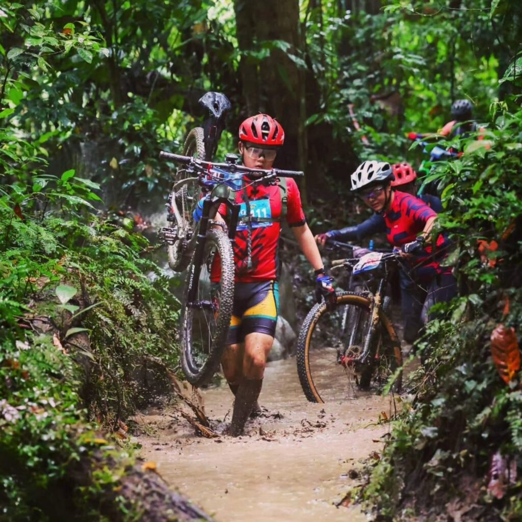 A line of mountain bikers carrying their bikes on their shoulder to tackle a stream crossing